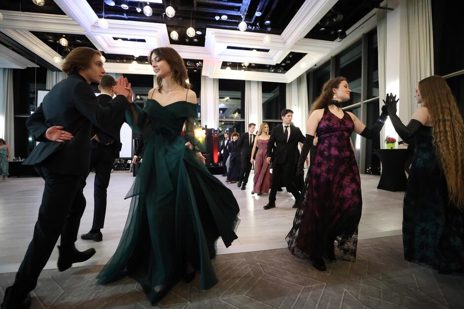 Kilka par wiruje na parkiecie. Są bardzo elegancko ubrani, kobiety mają wieczorowe długie suknie z odsłoniętymi ramionami, a mężczyźni garnitury.