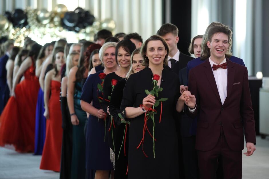 Grupa osób bardzo elegancko ubranych tańczy poloneza. W pierwszym rzędzie roześmiana kobieta trzyma w ręku czerwoną różę, jej partner też śmieje się szeroko. Ma na sobie garnitur i muchę.