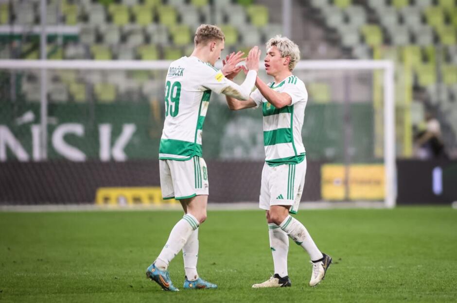 Dwaj piłkarze w biało-zielonych strojach stoją na boisku, przybijają piątkę po udanej akcji