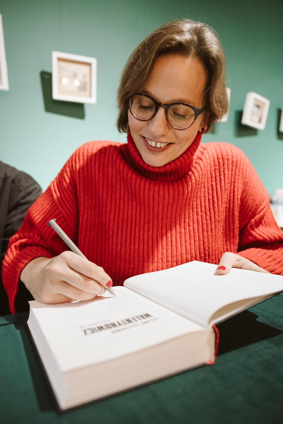 Młoda kobieta w okularach pochyla się nad książką. Trzyma w ręku długopis i pisze coś na pierwszej stronie książki. Jest ubrana w czerwony golf i uśmiecha się.