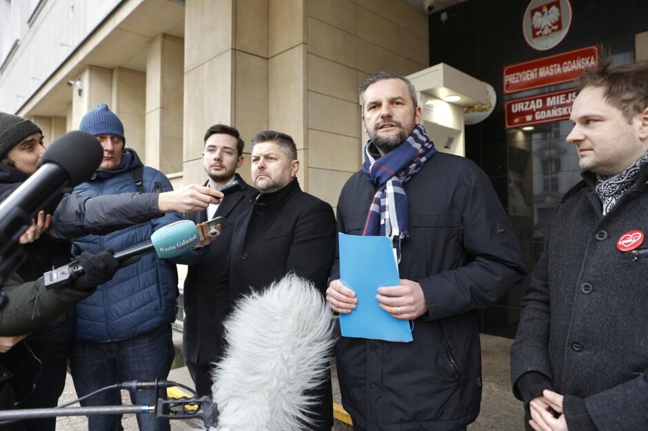 Na zdjęciu widzimy grupę mężczyzn stojących przed wejściem do budynku z napisem PREZYDENT MIASTA GDAŃSKA  oraz  URZĄD MIEJSKI w GDAŃSKU . Czworo z nich jest zwróconych w stronę mikrofonów, które trzymają reporterzy, sugerując, że udzielają wywiadu lub komentarza. Mężczyźni są ubrani w zimowe okrycia; jeden trzyma niebieską teczkę, a inny ma na piersi przypiętą czerwoną przypinkę z sercem. Scena ta prawdopodobnie przedstawia jakieś wydarzenie lub konferencję prasową na zewnątrz urzędu miejskiego. Atmosfera wydaje się być oficjalna i poważna
