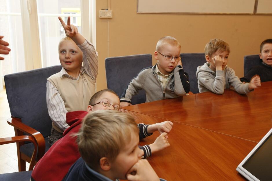 Kilkoro 6-latków siedzi przy stole, jeden podnosi rękę, zgłaszając, że chce coś powiedzieć