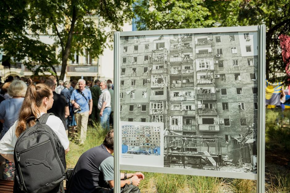 zdjęcie, na którym fragment skweru i ludzie oraz prezentowane zdjęcie - zbombardowanego budynku