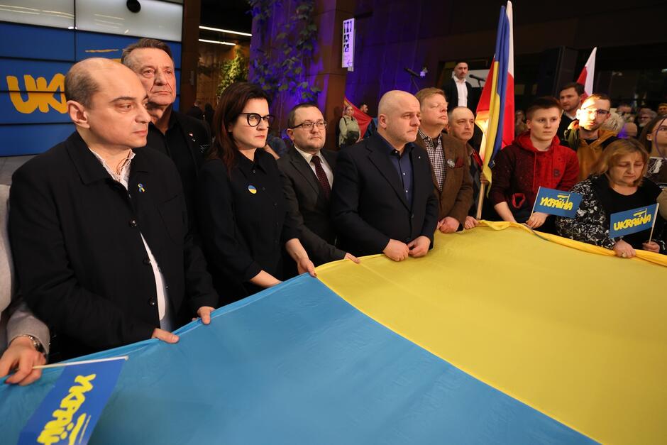 Na zdjęciu widać grupę osób trzymających duży, poziomy ukraiński flagę, na której dominują kolory niebieski i żółty. Osoby na zdjęciu mają różne wyrazy twarzy, od skupienia po powagę, co może sugerować, że uczestniczą w jakimś ważnym, być może politycznym lub solidarnościowym wydarzeniu. Niektórzy mają na sobie symbole lub przypinki, które mogą wyrażać ich wsparcie dla Ukrainy. Tło i oświetlenie wskazują, że są w środku, prawdopodobnie w budynku publicznym lub centrum konferencyjnym. Atmosfera wydaje się być formalna i zaangażowana