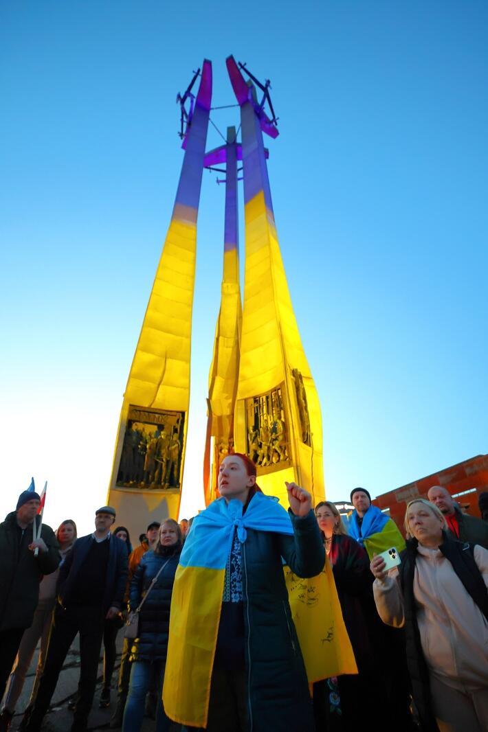 Na zdjęciu znajduje się grupa ludzi zebrana na zewnątrz wokół wysokiego pomnika, oświetlonego na żółto-niebiesko, w barwach narodowych Ukrainy. Na pierwszym planie widać kobietę owiniętą w żółto-niebieski szal, która podnosi rękę w geście, który może wyrażać solidarność lub protest. Tłum wygląda na zaangażowany, niektórzy patrzą w górę w kierunku flag lub konstrukcji. Sceneria i oświetlenie sugerują, że jest to wydarzenie wieczorne lub o zachodzie słońca, co dodaje dramatyzmu ujęciu. Niebo jest jasne i bezchmurne. Wydarzenie to prawdopodobnie ma charakter polityczny lub pamięciowy, co sugeruje wykorzystanie symboli narodowych i poważne wyrazy twarzy uczestników