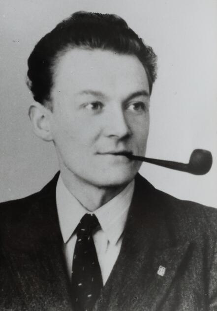 Młody mężczyzna w garniturze i białej koszuli z krawatem. Ma dość krótkie, ułożone do tyłu włosy i fajkę w ustach. Zdjęcie jest czarno-białe