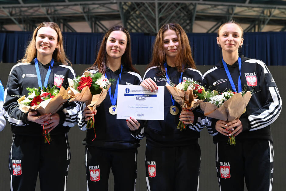 Cztery młode uśmiechnięte dziewczyny w dresach, w dłoniach trzymają wiązanki kwiatów, na szyjach wiszą medale