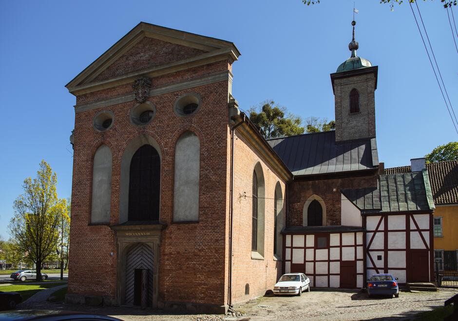 Na zdjęciu widać budynek o cechach kościoła, wykonany głównie z cegły, z wyraźnymi elementami architektonicznymi. Część frontowa ma prostą, ale elegancką fasadę z arkadowymi oknami i centralnie umieszczonym wejściem, nad którym widnieje data MDCLXVIII  (1668). Na boku kościoła znajduje się niższa, przylegająca konstrukcja z charakterystyczną, drewnianą, ryglową ścianą w białym i brązowym kolorze. Dachy budynku pokryte są blachą, co nadaje kontrastu czerwonej cegle. Za budynkiem kościoła widać wieżę z małym okienkiem i zielonym dachem zakończonym niewielką kopułą i krzyżem. W tle widoczne są drzewa i niebo bezchmurne, co wskazuje na ładną pogodę. Obok kościoła zaparkowane są dwa samochody, co wskazuje na współczesne ujęcie. Całość kompozycji znajduje się w miejskim lub podmiejskim otoczeniu, biorąc pod uwagę widoczne w oddali budynki mieszkalne