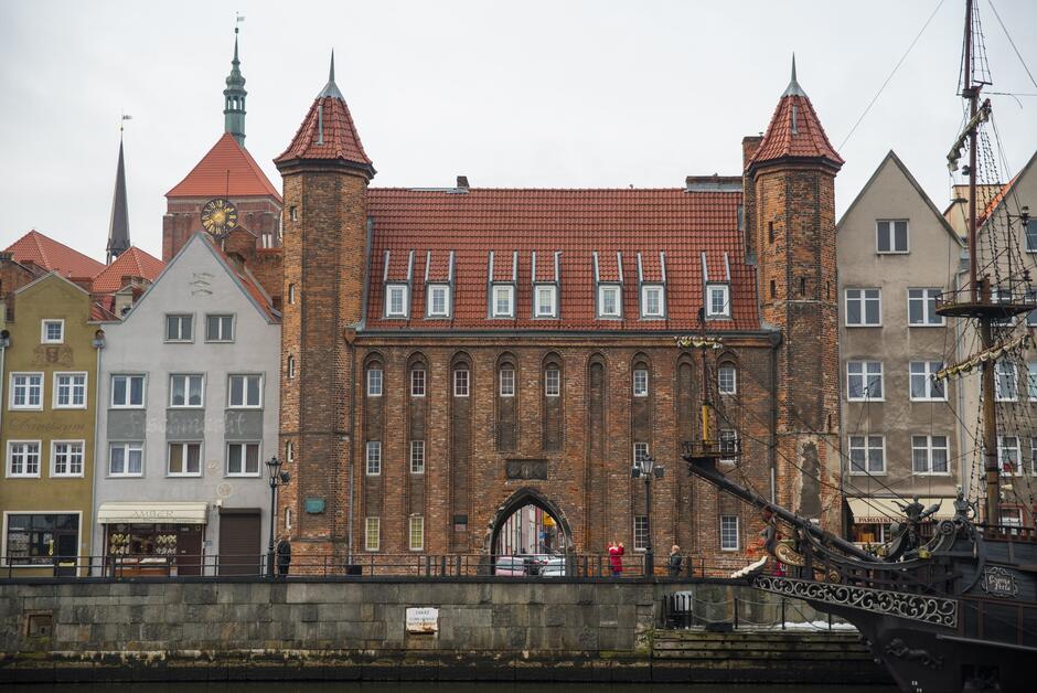Zdjęcie przedstawia fragment miasta z zabytkowymi budynkami o cechach europejskiej architektury. Centralny budynek ma charakterystyczne czerwone dachy i kilka szpiczastych wieżyczek, co nadaje mu wygląd typowy dla północnoeuropejskiego gotyku. Jego fasada jest wykonana z cegły, a w centralnej części widoczny jest duży łukowy przejazd. Po lewej stronie znajduje się węższy budynek z wyraźnie starszą fasadą i inskrypcją Schifffahrt . Poniżej, na poziomie chodnika, widoczne są witryny sklepowe. Po prawej stronie zdjęcia znajduje się kolejny budynek mieszkalny. Na pierwszym planie widać nabrzeże, przy którym cumuje żaglowiec, z którego dziób wyróżnia się dzięki bogato zdobionej rzeźbie. Na brzegu i nad wodą znajduje się kilka postaci ludzkich, co sugeruje, że jest to popularne miejsce spacerów lub turystyczny rejon miasta. Scena jest uchwycona w dzień, przy pochmurnym niebie, co nadaje zdjęciu nieco ponury nastrój, ale równocześnie podkreśla historyczny charakter środowiska.