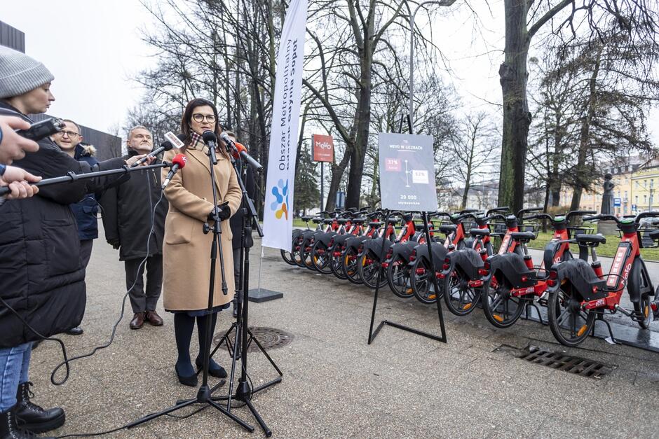 na zdjęciu kobieta w średnim wieku, w piaskowym płaszczu, mówi do dziennikarskich mikrofonów, po prawej stoi kilkanaście czerwonych rowerów, jeden obok drugiego
