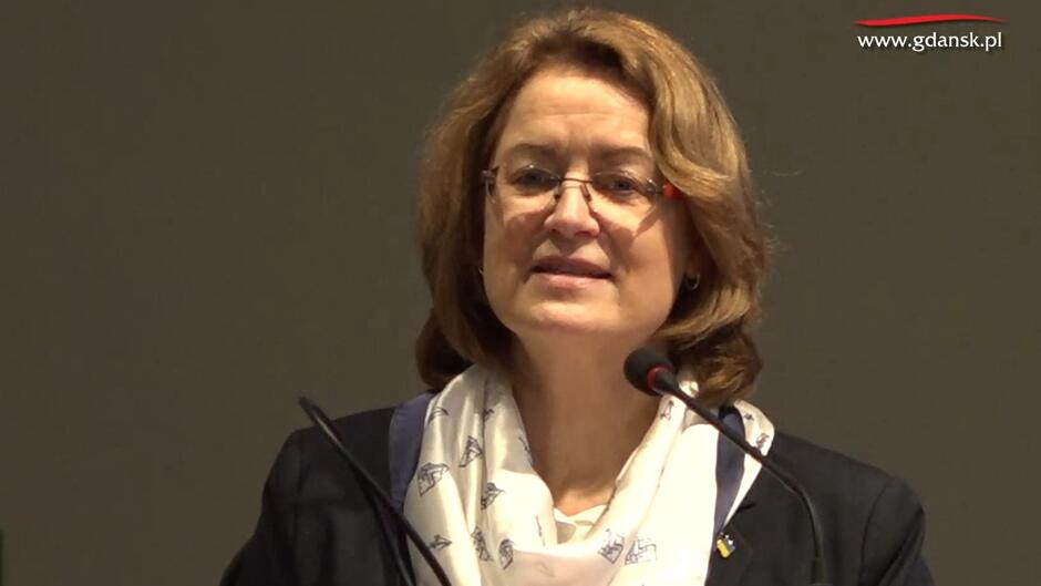 Kobieta w średnim wieku, o pofalowanych włosach, w okularach - jest w trakcie przemówienia