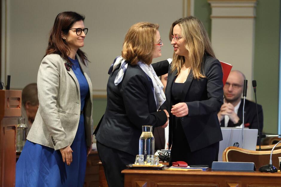 Trzy kobiety stoją obok siebie. Dwie po prawej są w trakcie przyjaznego uścisku na powitanie