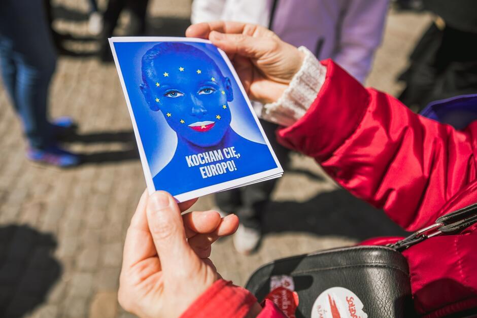 Zdjęcie przedstawia dwie dłonie, które trzymają okolicznościową kartę wielkości pocztówki. Widnieje na niej niebieska twarz i napis Kocham Cię Europo. Na niebieskiej twarzy umieszczony jest wianek żółtych gwiazdek symbolizujących UE, wargi ust kobiety mają kolory biały i czerwony