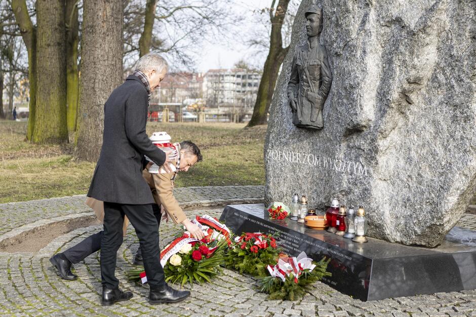 Dwie osoby podchodzą z kwiatami do kamiennego obelisku