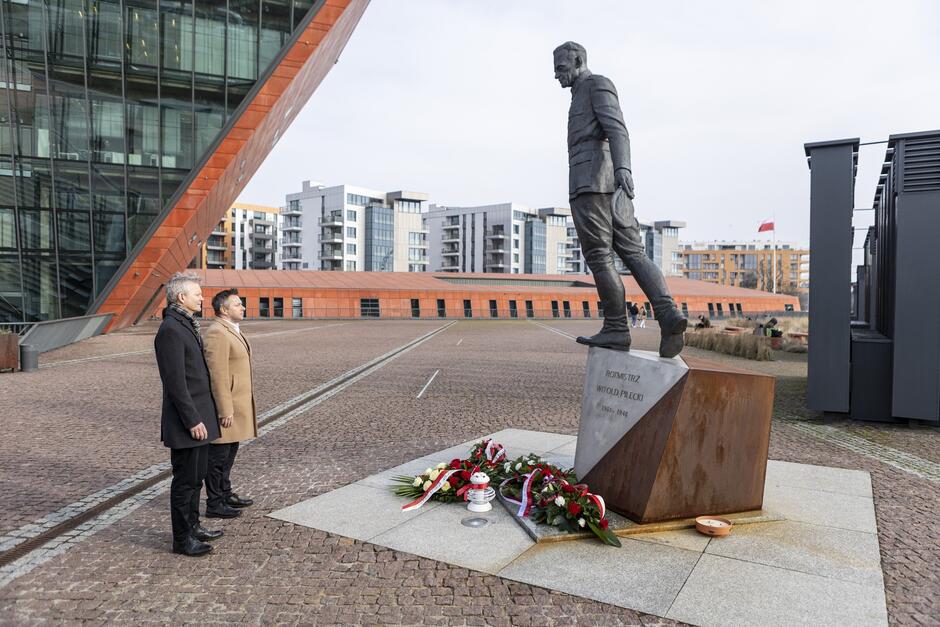 Pomnik przedstawia figurę idącego mężczyzny. Przed pomnikiem stoją dwaj panowie z kwiatami