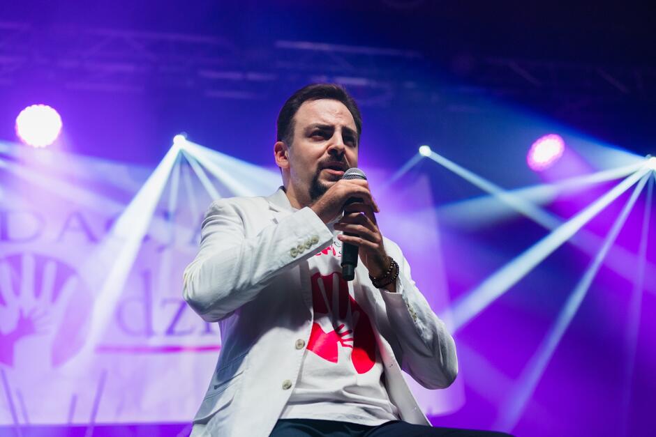 Mężczyzna w białej marynarce na scenie z mikrofonem. Siedzi na krześle