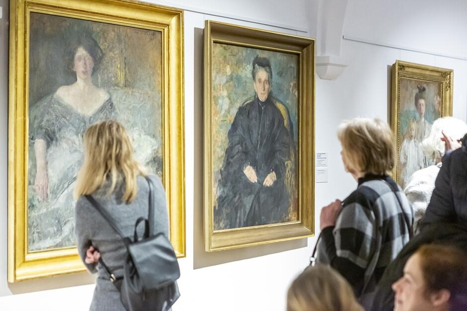 muzeum, przechodzący oglądają duże portrety zawieszone na ścianach w złotych ramach