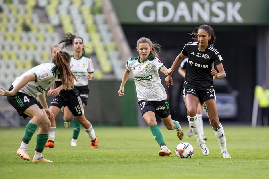 Po prawej przy piłce wysoka śniada zawodniczka w czarnym stroju, po lewej trzy niższe w białych koszulkach