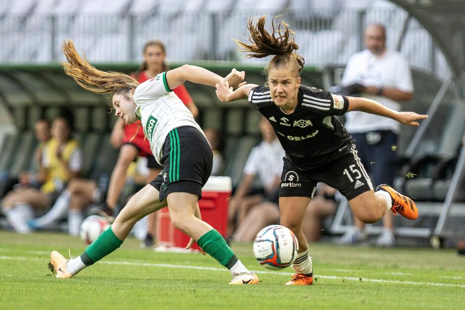 Dwie piłkarki: po lewej w białej koszulce, po prawej w czarnej walczą o piłkę, obie przechylone w lewo i prawo