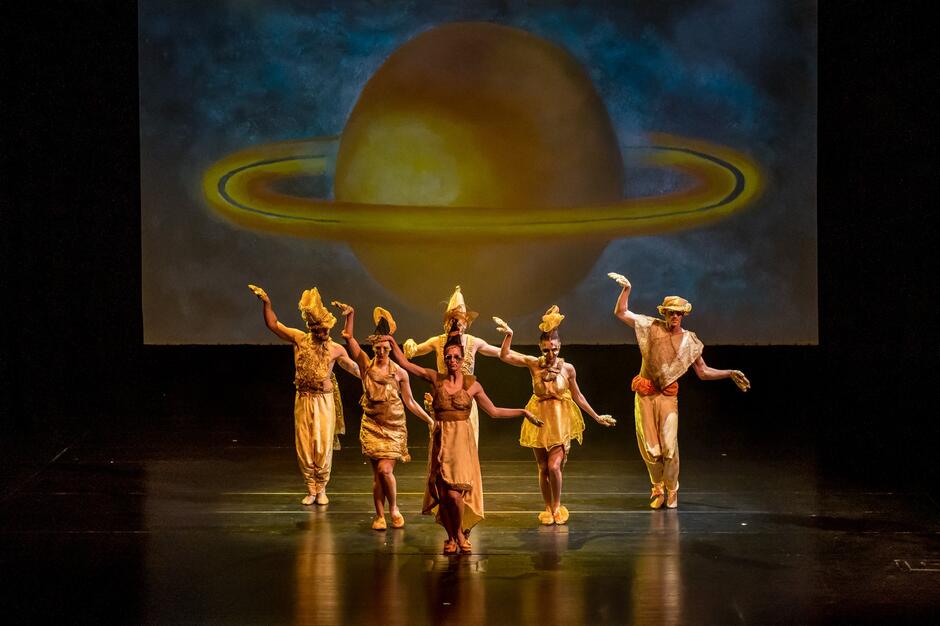 Kilka osób w pozłacanych strojach. Za nimi na ścianie wizerunek planety Saturn