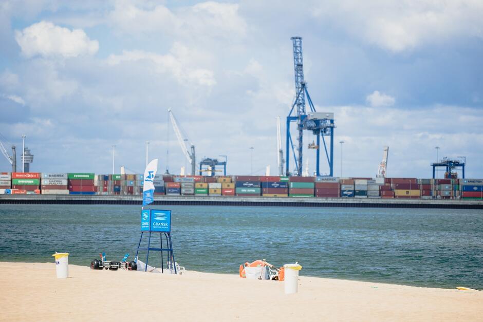 na zdjęciu plaża, widać na niej kilka kubłów na śmieci, w tle kontenerowce z dźwigiem portowym