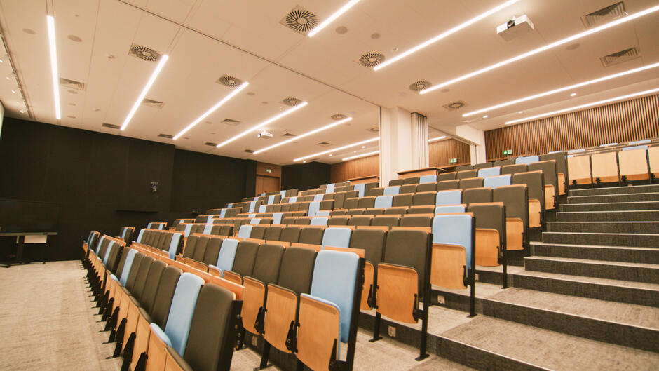 To zdjęcie pokazuje wnętrze nowoczesnej auli wykładowej lub sali konferencyjnej. Widoczne są rzędy ułożonych amfiteatralnie krzeseł z tapicerką w ciepłych odcieniach brązu i niebieskiego, które zapewniają komfortowe siedzenie. Sala jest dobrze oświetlona dzięki szeregom równo rozmieszczonych świetlówek na suficie, które zapewniają równomiernie rozproszone światło. Na suficie zamontowane są również okrągłe kratki wentylacyjne. Przód sali zdobi czarna ściana, obok której stoi biały ekran lub tablica interaktywna, a na ścianach bocznych widać dekoracyjne panele drewniane. Całość sprawia wrażenie nowoczesnego i funkcjonalnego miejsca przeznaczonego do nauki lub prezentacji