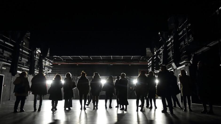 Kilkanaście osób stoi na krawędzi sceny, widzimy zarys ich sylwetek od tyłu, są oświetlone światłem rampy. Są zwróceni twarzami do widowni. Zdjęcie jest czarno-białe.