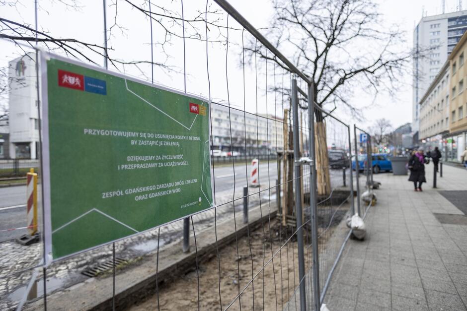 na zdjęciu fragment siatki ochronnej odgradzającej plac budowy z zieloną tablicą informacyjną o prowadzonych pracach