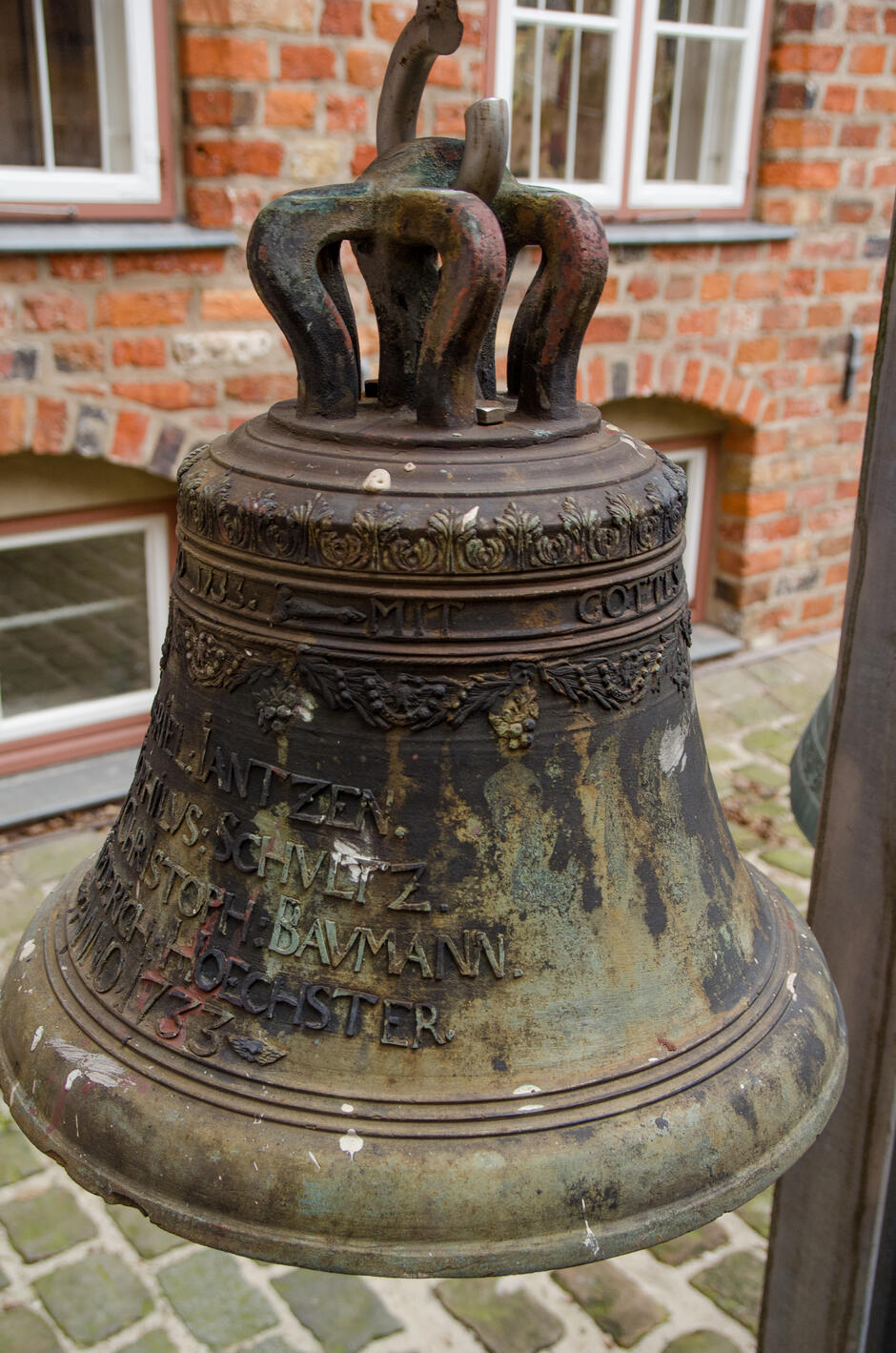To zdjęcie przedstawia stary dzwon z brązu z różnymi inskrypcjami, umieszczony na zewnątrz obok ceglanej ściany. Na górze dzwonu znajduje się dekoracyjny pas z liściastym wzorem, a poniżej niego inskrypcje wypukłymi literami. Można wyróżnić słowa MIT GOTT , a także kilka imion i nazwisk, takich jak  CHRISTOPH BAUMANN , oraz datę  1750 . Dzwon ma tradycyjny kształt, z widocznym uchwytami na górze. Powierzchnia dzwonu wykazuje oznaki korozji i patynowania, z zielonkawymi przebarwieniami, co sugeruje jego starość i długotrwałe wystawienie na działanie czynników atmosferycznych