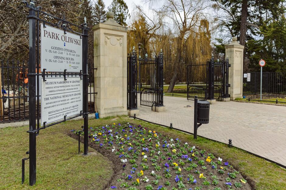 na zdjęciu brama wejściowa do parku, w tle jesienne drzewa bez liści, na pierwszym planie stylizowana tablica informacyjna z nazwą park oliwski