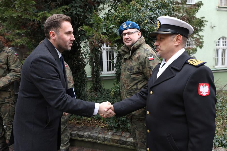 Mężczyzna w cywilnym ubiorze stoi po lewej, podaje rękę mężczyźnie w oficerskim mundurze marynarskim, który stoi po prawej. W głębi stoi oficer w zielonym mundurze i niebieskim berecie