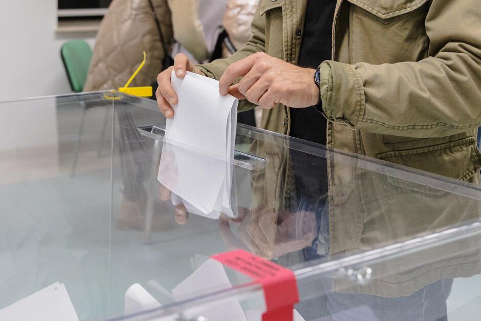 Męska ręka w kurtce wrzuca kartkę do przezroczystej urny wyborczej