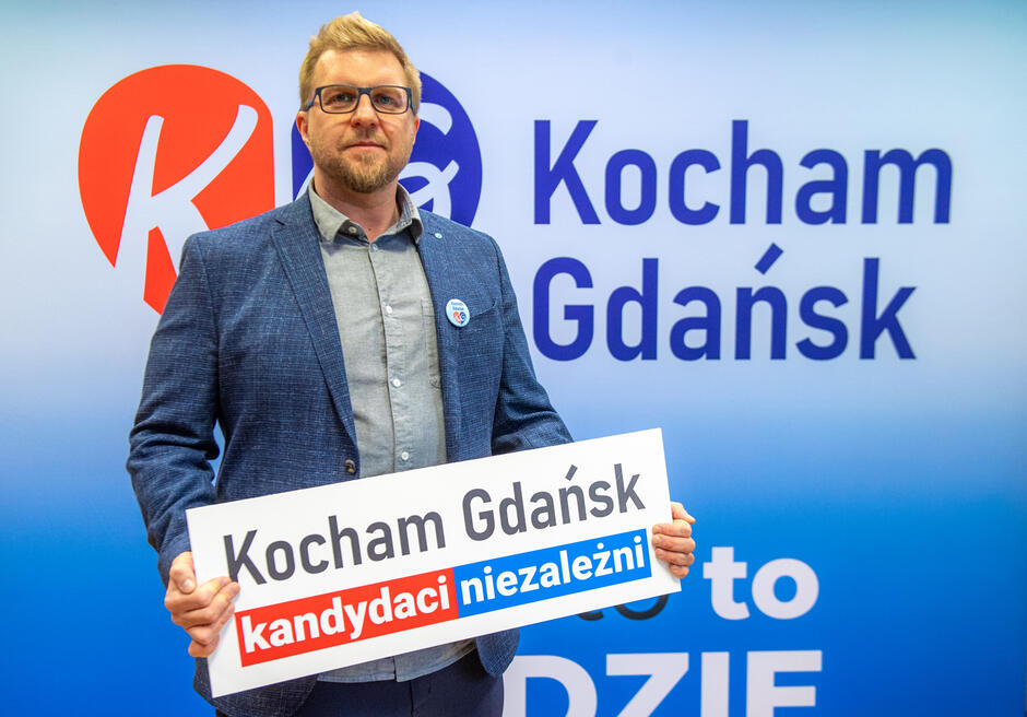 Mężczyzna w okularach i niebieskim garniturze trzyma tabliczkę z napisem Kocham Gdańsk kandydaci niezależni  przed tłem z logo i hasłem  Kocham Gdańsk  na niebieskiej ściance
