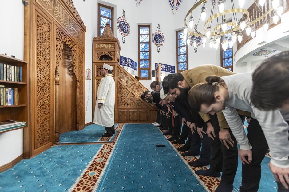 na zdjęciu pochyleni mężczyźni, którzy stoją w rzędzie jeden obok drugiego, przed nimi stoi wyprostowany duchowny, imam w białej długiej szacie