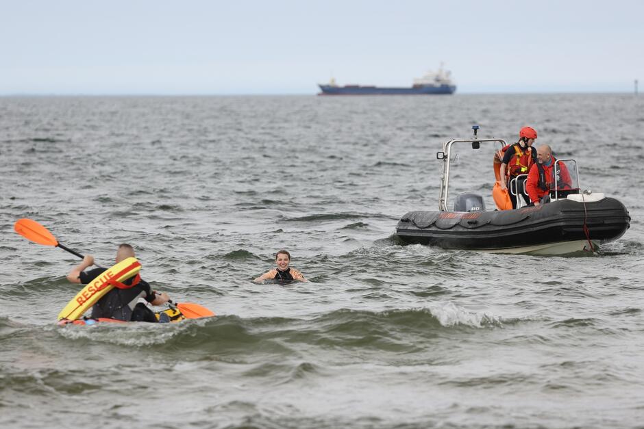 kobieta w wodzie udaje, że tonie, za nią na łodzi motorowej dwóch ratowników, z przodu na łodzi wiosłowej jeden