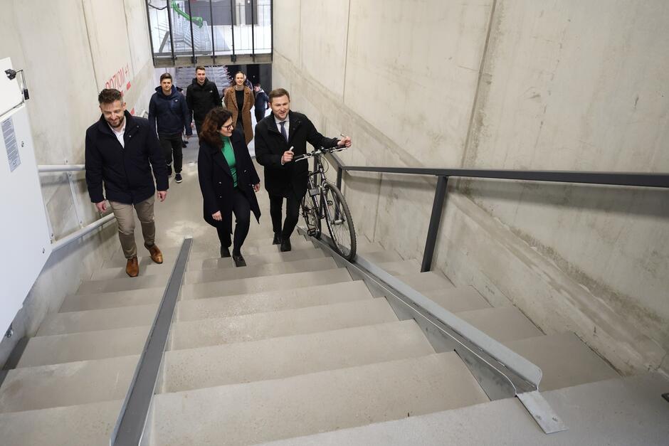 na zdjęciu kilka osób wchodzi po schodach, jeden z mężczyzn prowadzi rower, wjeżdża on na specjalnej metalowej platformie