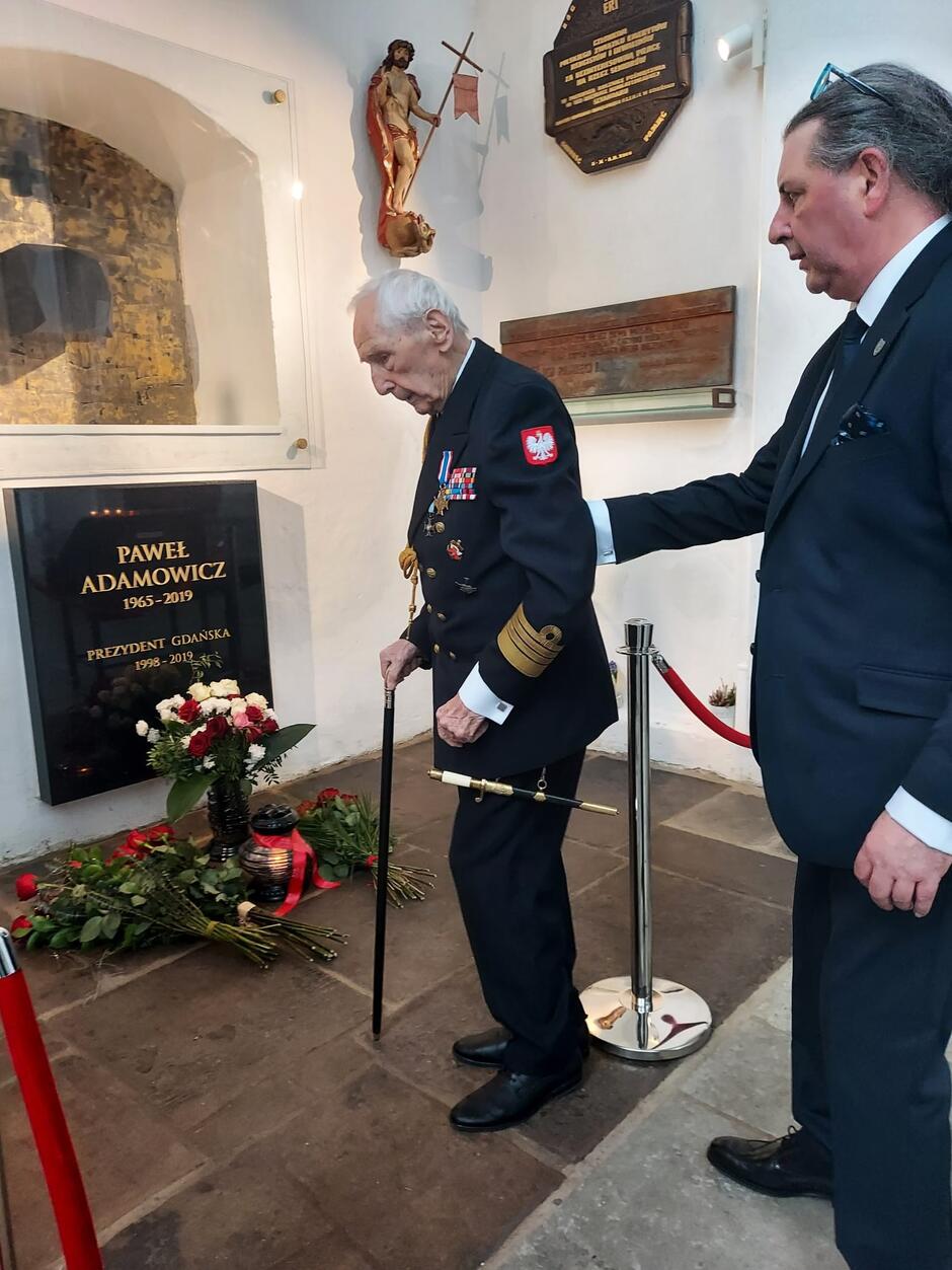 Mężczyzna w mundurze marynarki wojennej, stoi przed płytą w ścianie kościoła, na płycie jest napis Paweł Adamowicz. Przed płyta, na posadzce, leżą kwiaty