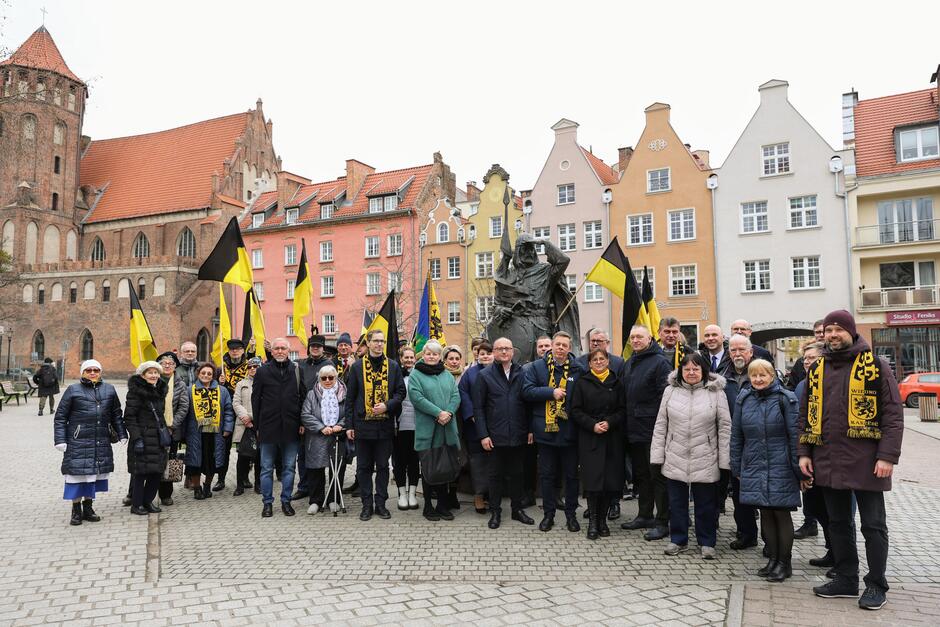 Kilkadziesiąt osób kobiet i mężczyzn w różnym wieku w kurtkach i płaszczach stoi przed pomnikiem rycerza. Niektórzy mają na szyi czarno-żółte szaliki. Z tyłu kilkupiętrowe kamienice  