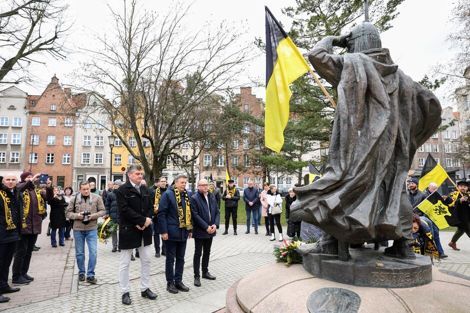 Z prawej strony fragment pomnika rycerza, przed nim kilkoro osób, kobiet i mężczyzn w różnym wieku, trzymają w rękach wiązanki kwiatów