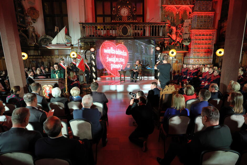 zdjęcie sali podczas uroczystości, na krzesłach siedzą goście, na scenie Jerzy Owsiak, obok przy mikrofonie stoi prezydentka Gdańska