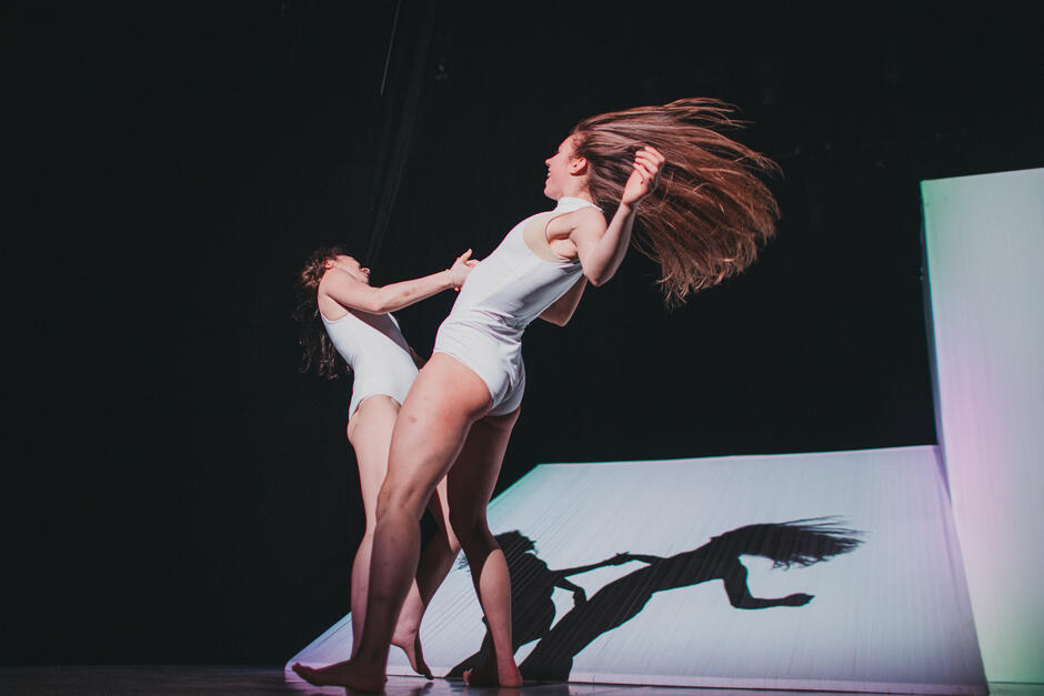 Dwie tancerki na scenie w pozycjach tanecznych, trzymają się za prawe ręce i odchylają do tyłu. Są ubrane w białe skąpe stroje, długie włosy jednej z nich unoszą się w ruchu.  