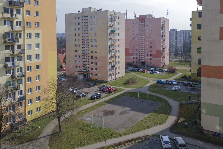 zdjęcie z drona, widać pięć wysokich bloków mieszkalnych, pomiędzy nimi zielony teren i asfaltowe spore boisko