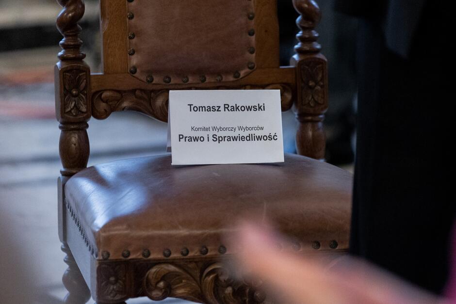 Na zdjęciu widać zbliżenie na drewniane krzesło z tabliczką, na której napisano Tomasz Rakowski Komitet Wyborczy Wyborców Prawo i Sprawiedliwość . Krzesło ma solidne drewniane ramiona z dekoracyjnymi rzeźbieniami i jest wykończone ćwiekami. Tabliczka z imieniem i nazwiskiem sugeruje, że krzesło jest zarezerwowane dla osoby o tym imieniu, prawdopodobnie uczestniczącej w jakimś wydarzeniu politycznym lub oficjalnym spotkaniu. Tło jest nieostrze, co pozwala się skupić na przedstawionym przedmiocie