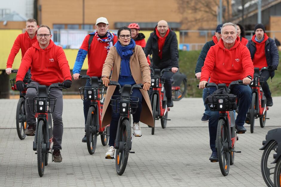 Grupa ludzi ubranych głównie w czerwone bluzy z logo „Mevo” jeżdżących na rowerach miejskich. Są ustawieni w dwóch rzędach, jeden za drugim, i poruszają się po płaskim, brukowanym placu przed Ergo Areną. Na czele grupy kobieta (prezydent Gdańska) w camelowym płaszczu i niebieskim szaliku 