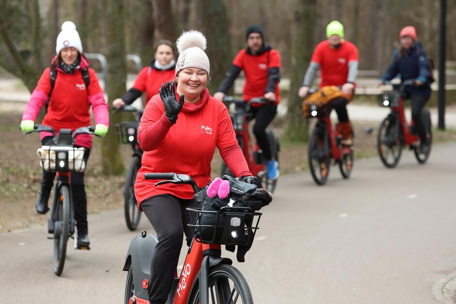 To zdjęcie przedstawia grupę ludzi na rowerach, którzy biorą udział w wspólnej przejażdżce lub wydarzeniu rowerowym. Osoba na pierwszym planie, kobieta w czerwonym ubraniu z rękawiczkami, macha do kamery z uśmiechem. Nosi ona białą czapkę i ma koszyk z przodu roweru. Pozostali uczestnicy wydarzenia rowerowego również są ubrani w czerwone i ciemne stroje, niektórzy z nich mają na głowach czapki w różnych kolorach.