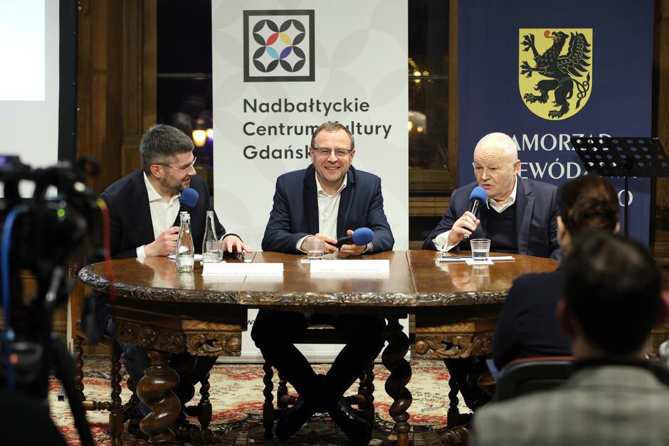 Na zdjęciu widać trzech mężczyzn siedzących przy okrągłym stole w trakcie jakiegoś wydarzenia lub panelu dyskusyjnego. Od lewej strony: pierwszy mężczyzna ma ciemne włosy, nosi ciemny garnitur i niebieską koszulę, wydaje się, że coś mówi lub wyjaśnia, patrząc na osobę pośrodku. Drugi mężczyzna, również w ciemnym garniturze, niebieskiej koszuli i okularach, uśmiecha się i trzyma mikrofon. Mężczyzna po prawej stronie, o łysiejącej głowie, nosi ciemny sweter z koszulą, trzyma mikrofon i wygląda na to, że zabiera głos. W tle widać baner z logotypem Nadbałtyckie Centrum Kultury Gdańsk  oraz herb. 