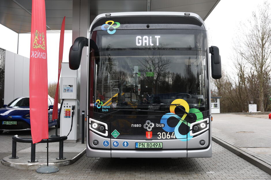 Widzimy przód czarnego autobusu elektrycznego, który jest podłączony do stacji ładowania. Na elektronicznym wyświetlaczu autobusu widnieje napis GAiT . Po obu stronach frontu autobusu umieszczone są grafiki i emblematy, w tym logo  neso bus  oraz symbol gazu ziemnego, informacje o dostępności dla osób niepełnosprawnych, a także herb i flaga Polski. Po lewej stronie autobusu znajduje się czerwona flaga z odwróconym herbem Danii. W tle, po lewej stronie, widać niebieski samochód sportowy, a za nim budynki przypominające tymczasowe biura na placu budowy. Wszystko to dzieje się w ciągu dnia i otoczenie wydaje się być częścią terenu komercyjnego lub przemysłowego