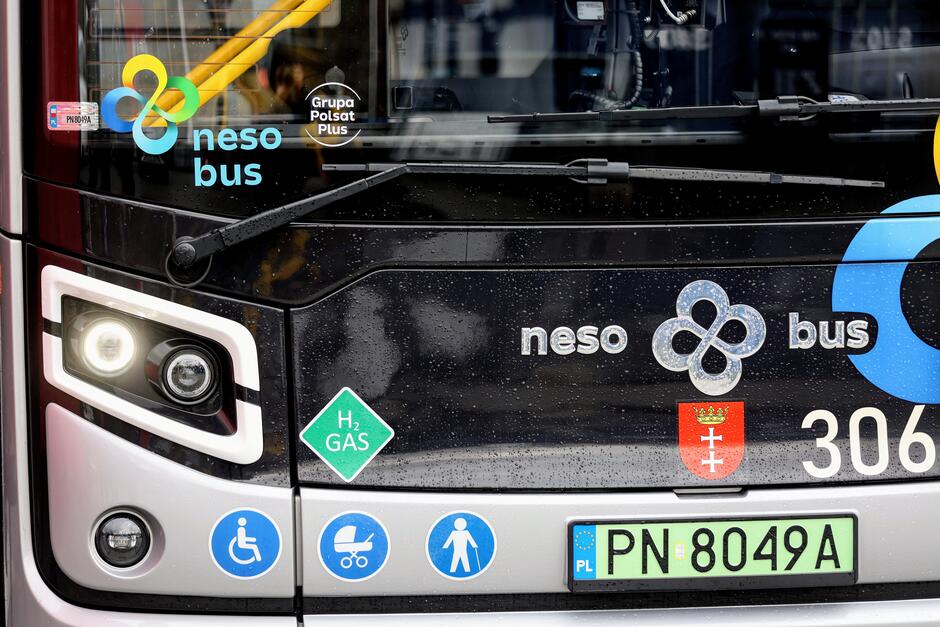 Zdjęcie przedstawia zbliżenie na przednią część czarnego autobusu z grafiką i napisami. Można zauważyć, że jest to autobus elektryczny z logotypem neso bus  oraz oznaczeniami dotyczącymi gazu wodorowego (H2 GAS). Ponadto na autobusie widnieje godło Polski oraz numery boczne  3064 . Na tablicy rejestracyjnej znajduje się oznaczenie  PN 8049A  z flagą Unii Europejskiej i inicjałami  PL , wskazującymi na polską rejestrację. Pod frontowymi światłami znajdują się symboliki dostępności, w tym dla osób niepełnosprawnych i z wózkami dziecięcymi. W tle przez szybę autobusu widać odbicie, co świadczy o tym, że zdjęcie zostało wykonane w ciągu dnia