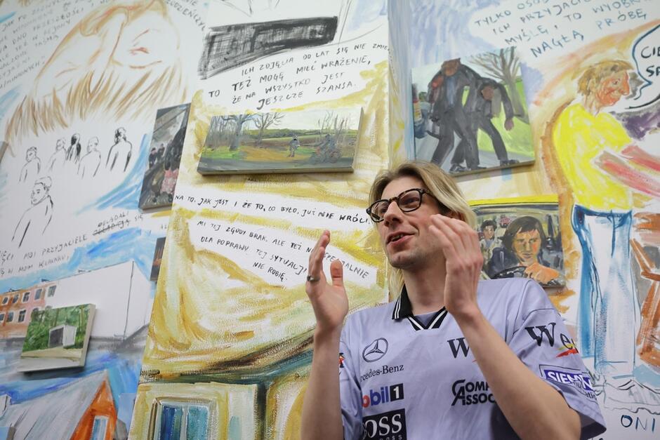 Młody mężczyzna w okularach i kolorowej koszuli na tle ściany wypełnionej napisami, rysunkami i obrazami. Podnosi ręce w gestykulacji, uśmiecha się