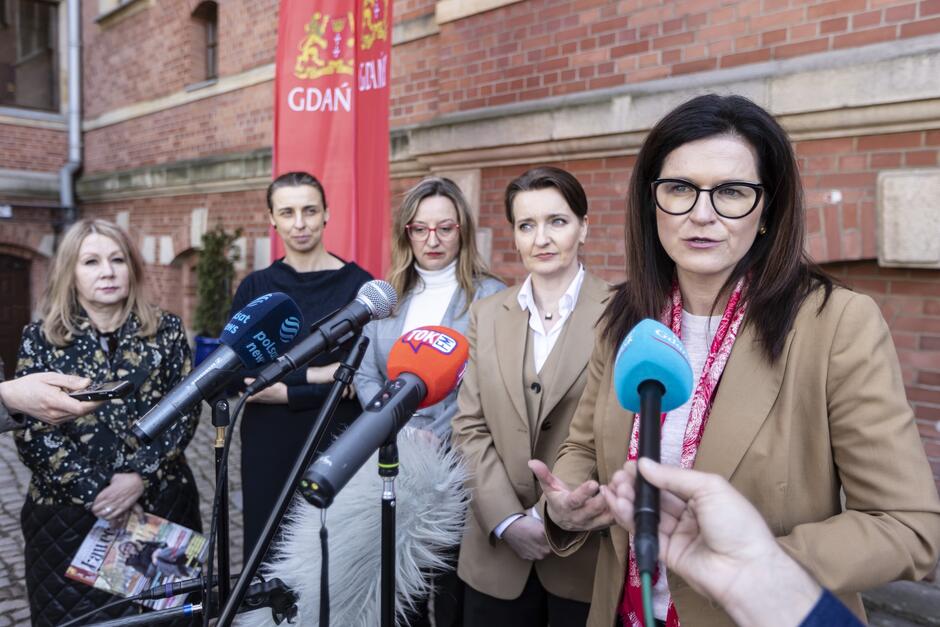 na zdjęciu pięć kobiet w oficjalnych strojach, pierwsza z nich mówi do mikrofonów dziennikarskich, pozostałe stoją obok w rzędzie